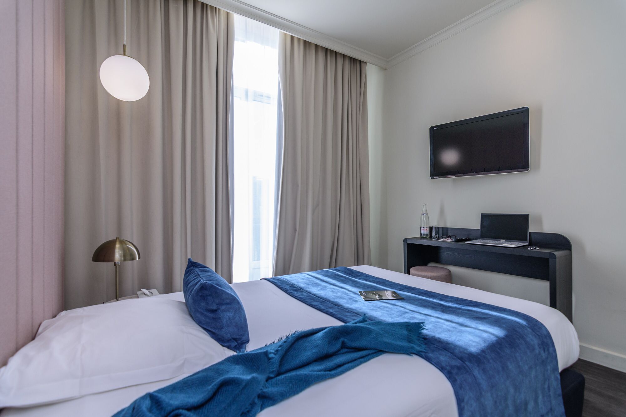 A modern hotel room with a large bed, a desk with a laptop, a flat-screen TV, and a large window with curtains.