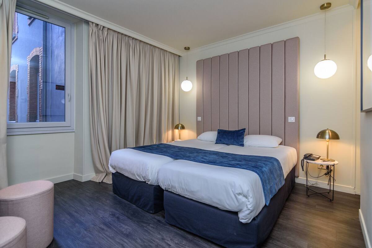 Habitación de hotel moderna con una cama grande, dos lámparas de noche, un ventanal con cortinas y vistas a un edificio.