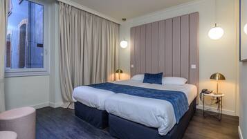 A modern hotel room with a large bed, two bedside lamps, a window with curtains, and a view of a building outside.