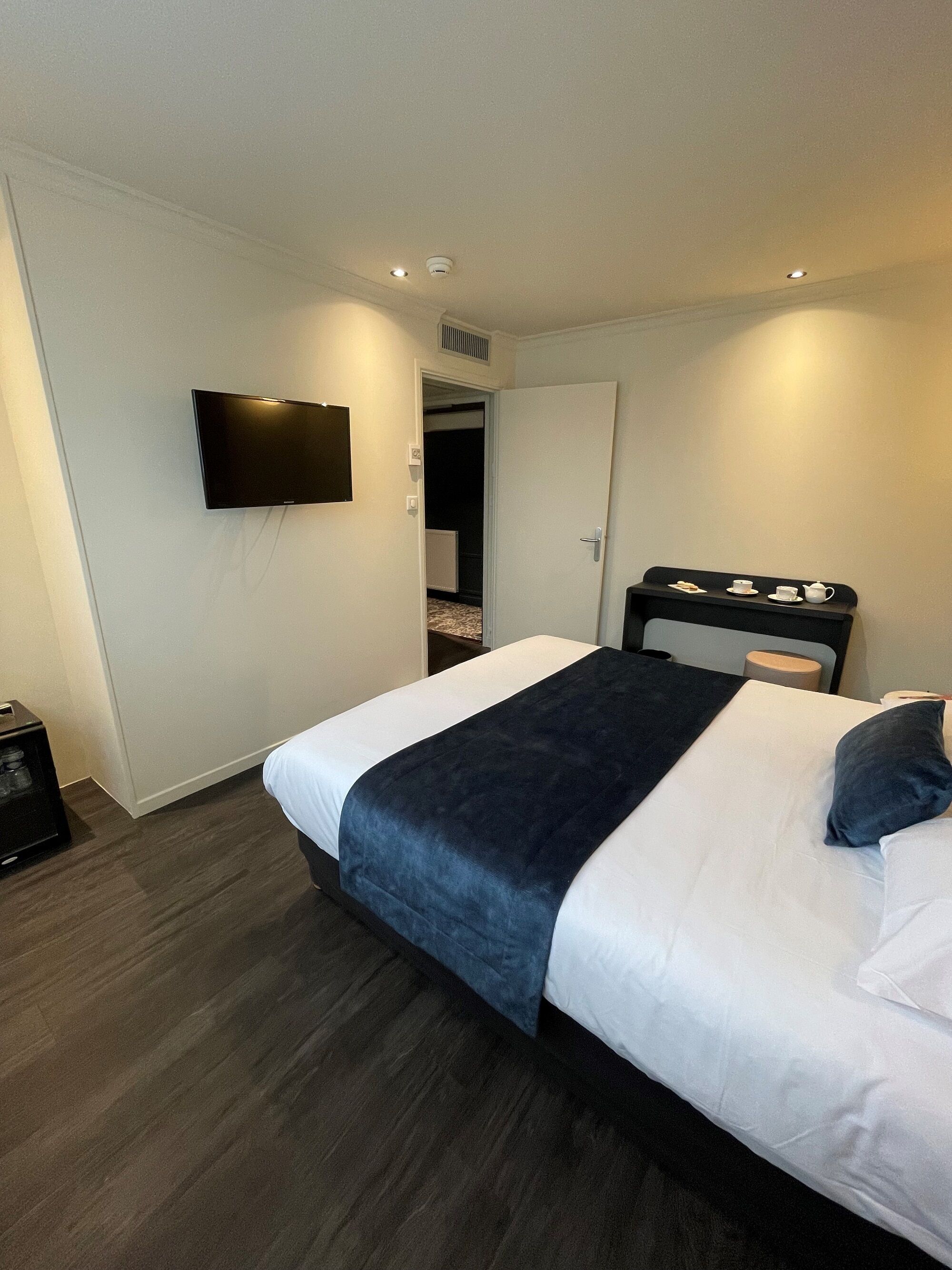 A hotel room with a bed, a flat-screen TV mounted on the wall, a bedside table with a tray, and a dark wooden floor.