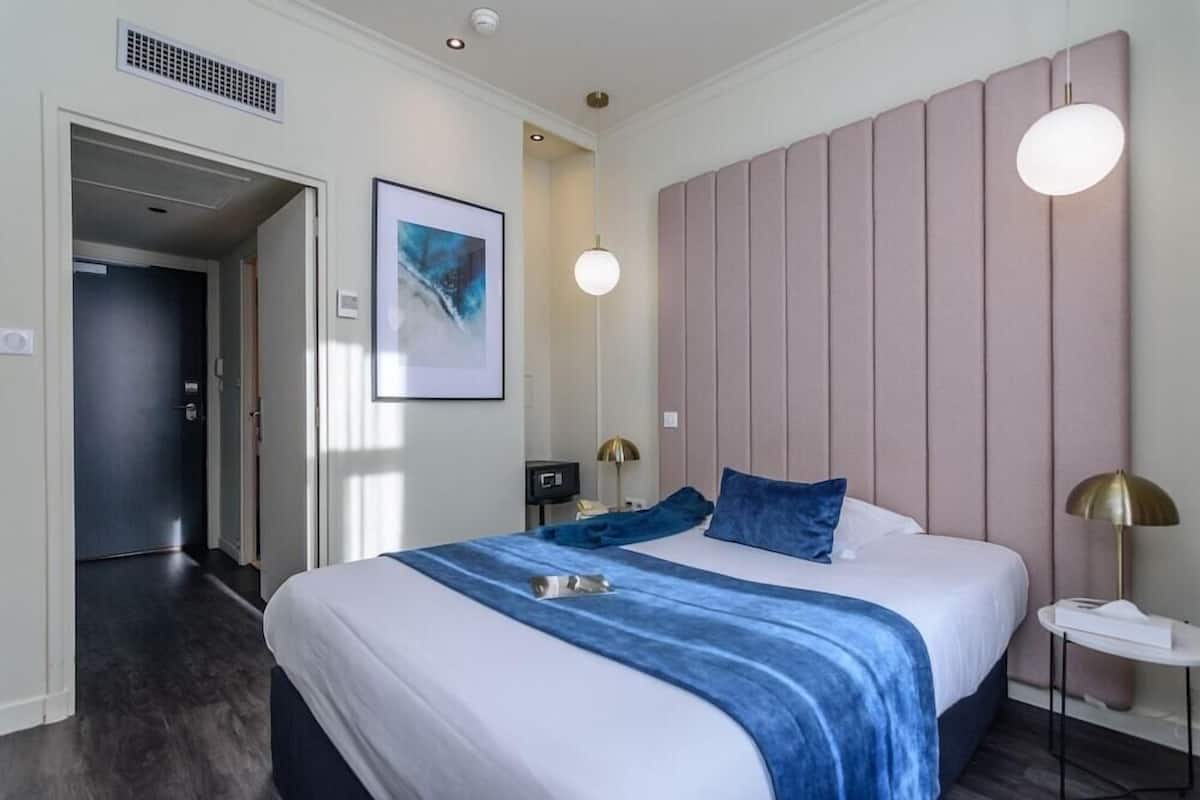 Habitación de hotel con una cama grande, una mesita de noche, una lámpara de pared y una puerta que da a otra habitación.