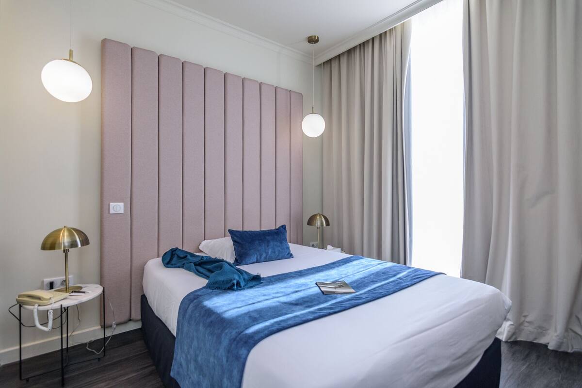 Una habitación de hotel con cama, lámparas de noche, teléfono y ventana con cortinas.