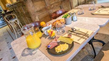 Un desayuno con una mesa de mármol, una cesta de mimbre con croissants, un plato de huevos revueltos y salami, y una jarra de vidrio con jugo de naranja.