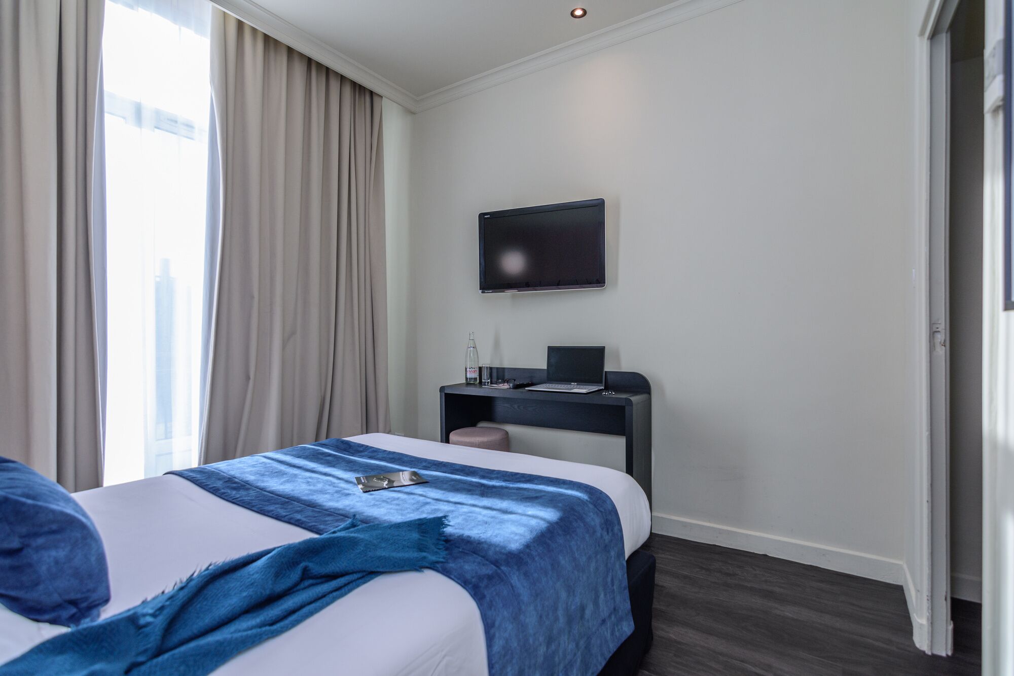 Una habitación de hotel moderna con una cama, un escritorio con una computadora portátil, un televisor y una ventana con cortinas.