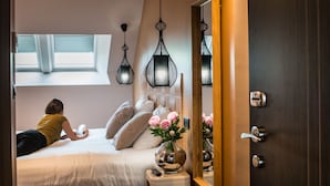 A bedroom with a bed, a bedside table, a vase with flowers, a mirror, and a window with blinds.