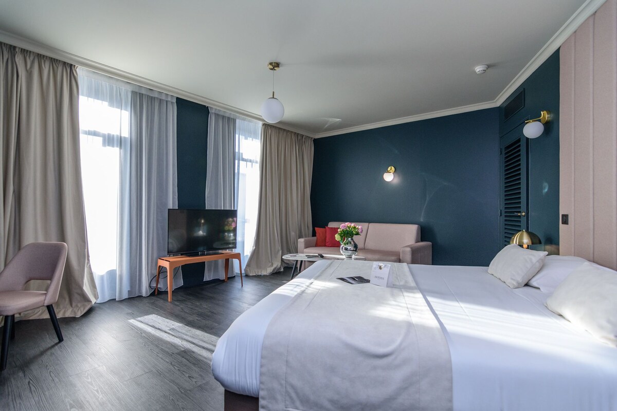 A hotel room with a bed, a TV on a stand, a sofa, a chair, and a small table with a vase.