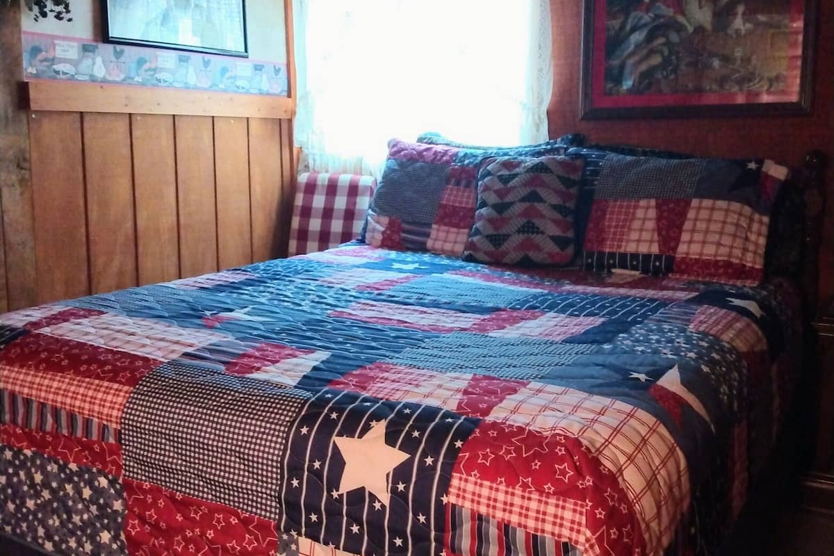 A bedroom with a large bed covered in a patchwork quilt, wooden walls, a window with curtains, and a framed picture on the wall.