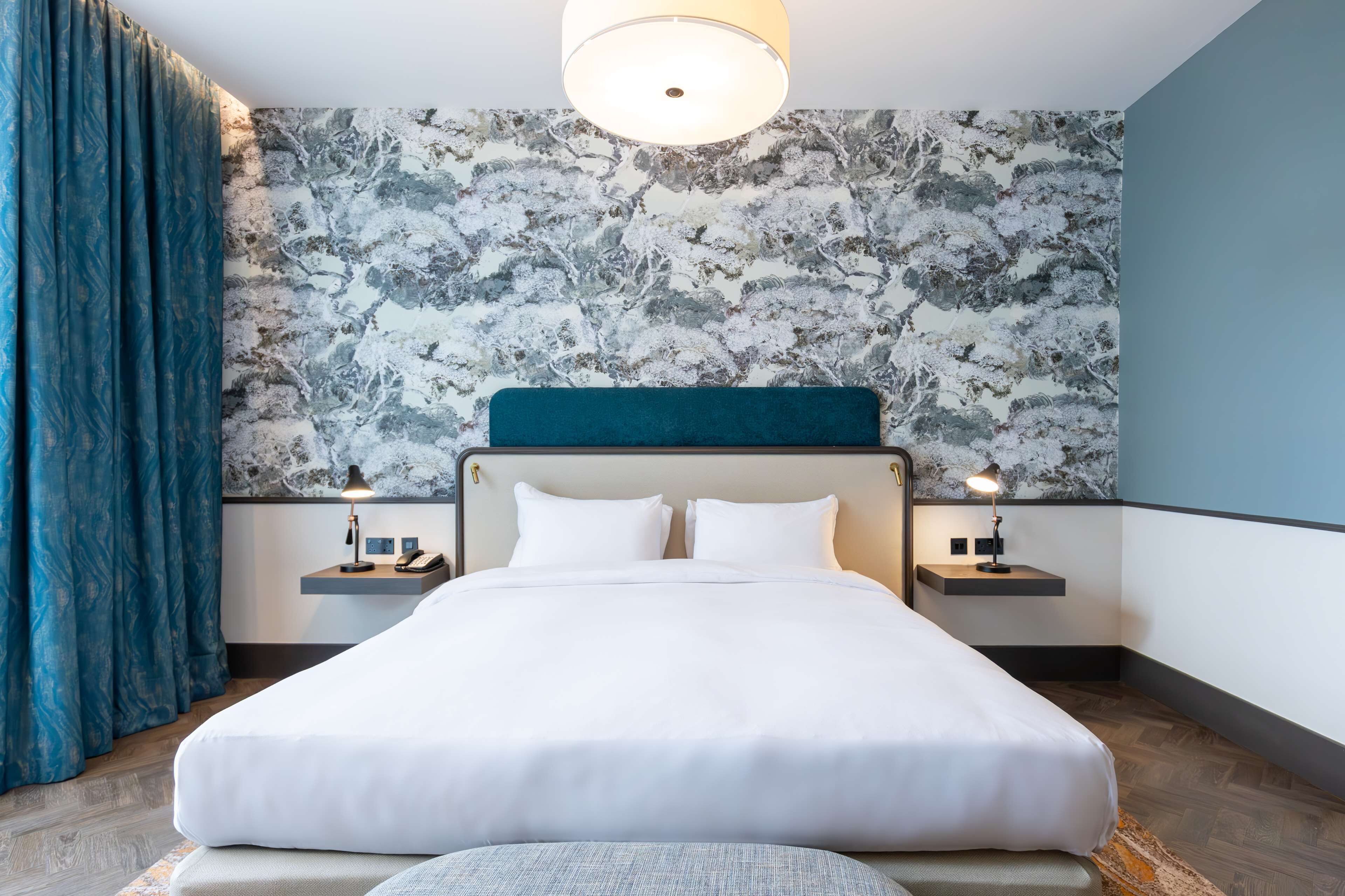 Habitación de hotel con una cama grande, dos mesitas de noche con lámparas, un cabecero y un papel tapiz estampado.
