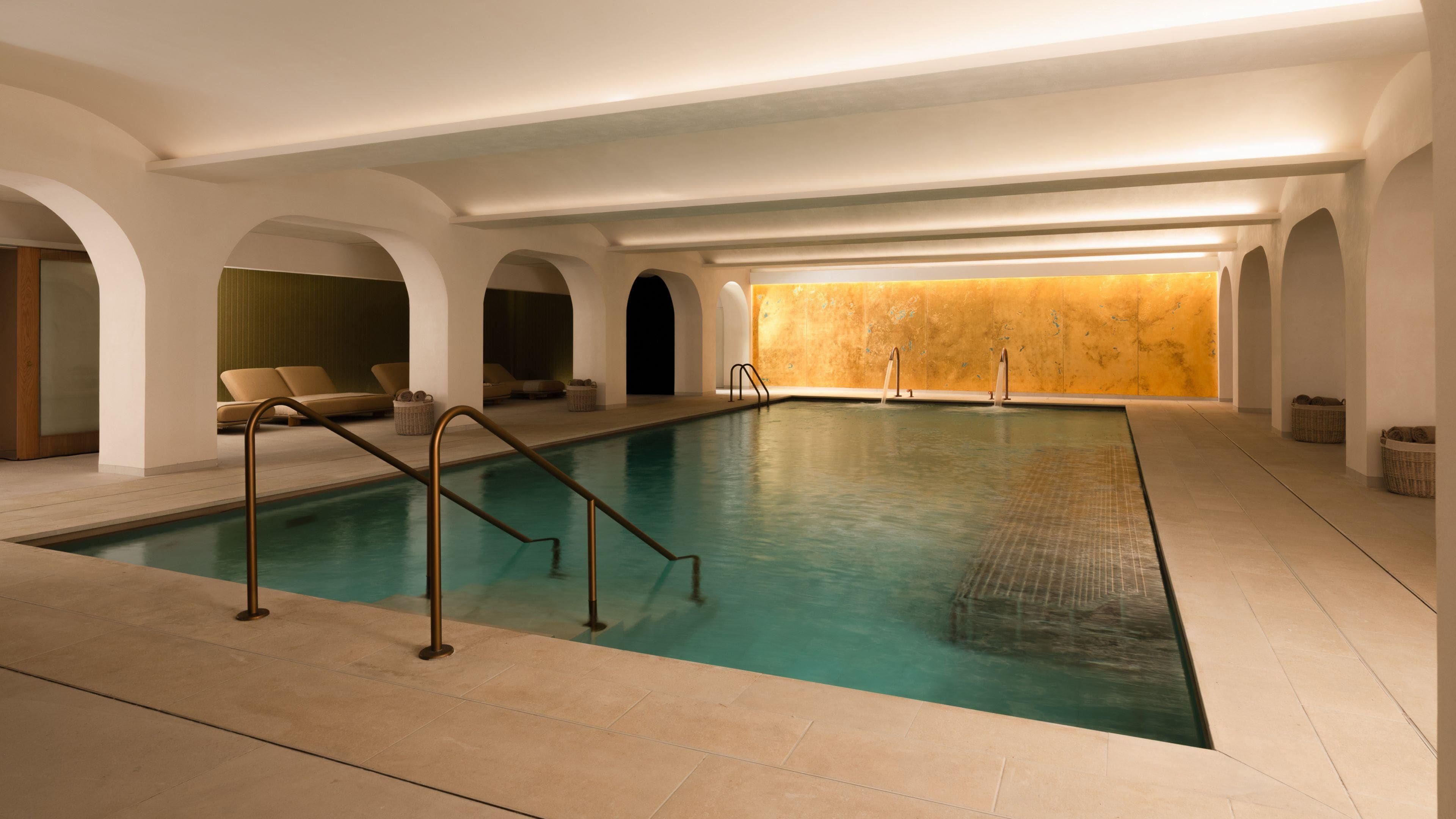 Una piscina con suelo de azulejos, rodeada de sillas de descanso y una pared con superficie texturizada.