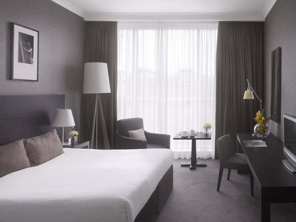 A hotel room with a large bed, a desk with a chair, a lamp, a TV, and a view of the city through sheer curtains.