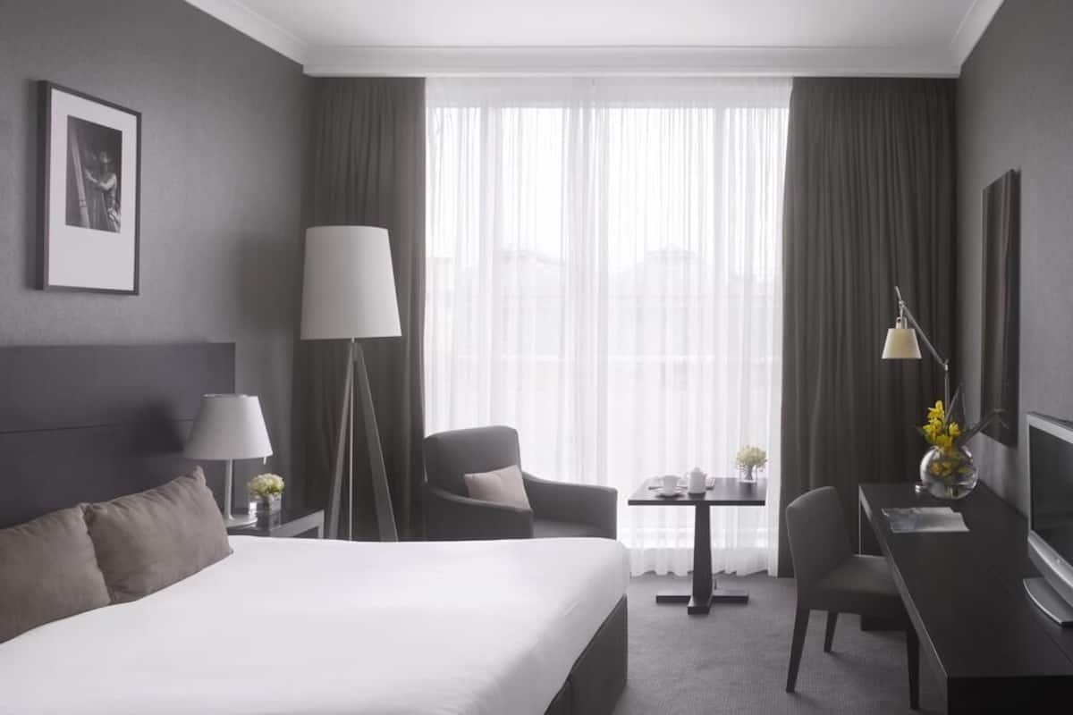 A hotel room with a large bed, a desk with a chair, a lamp, a TV, and a view of the city through sheer curtains.
