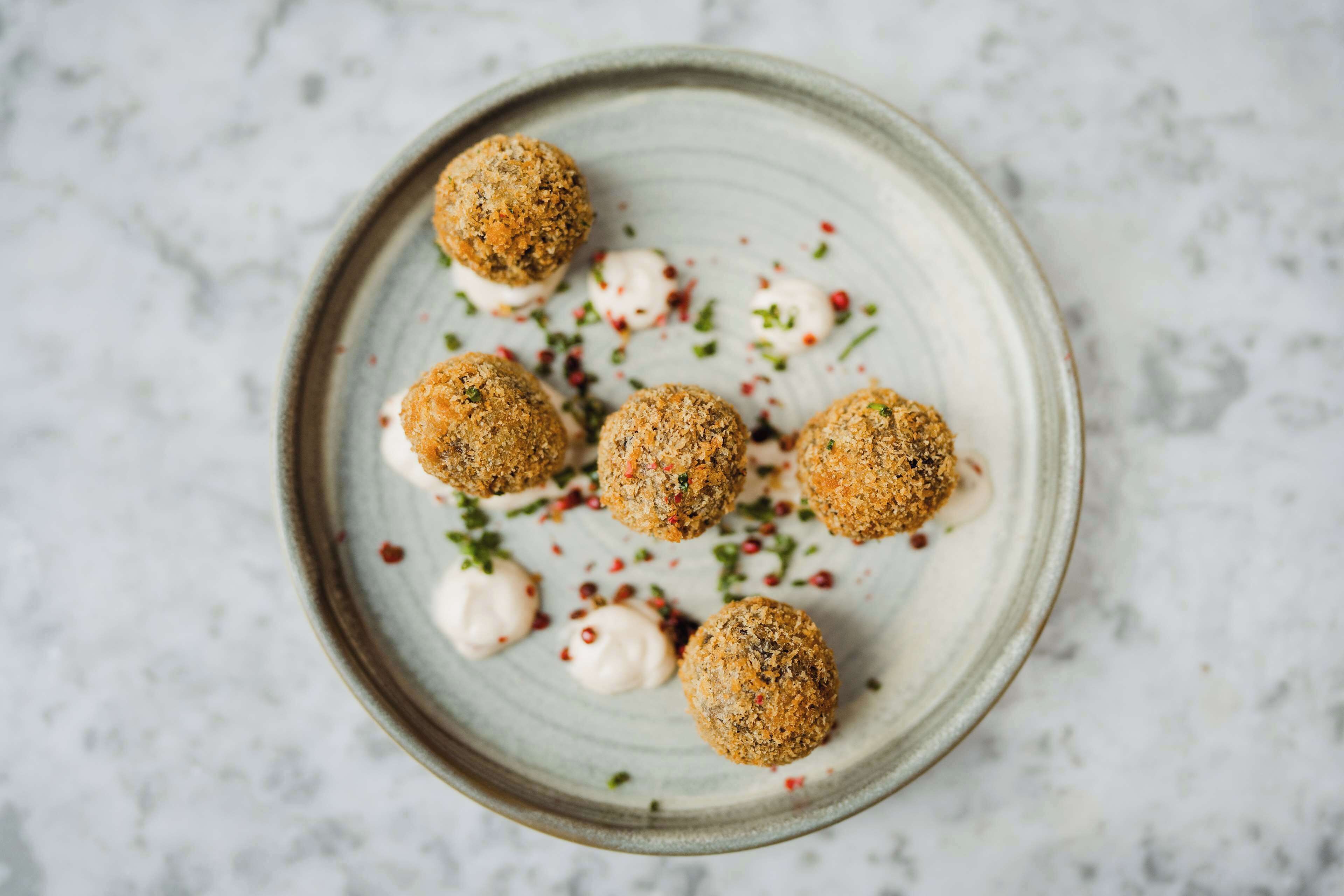 Un plato con seis bolas redondas rebozadas y cucharadas de salsa blanca, adornado con hojuelas de chile rojo y hierbas verdes.