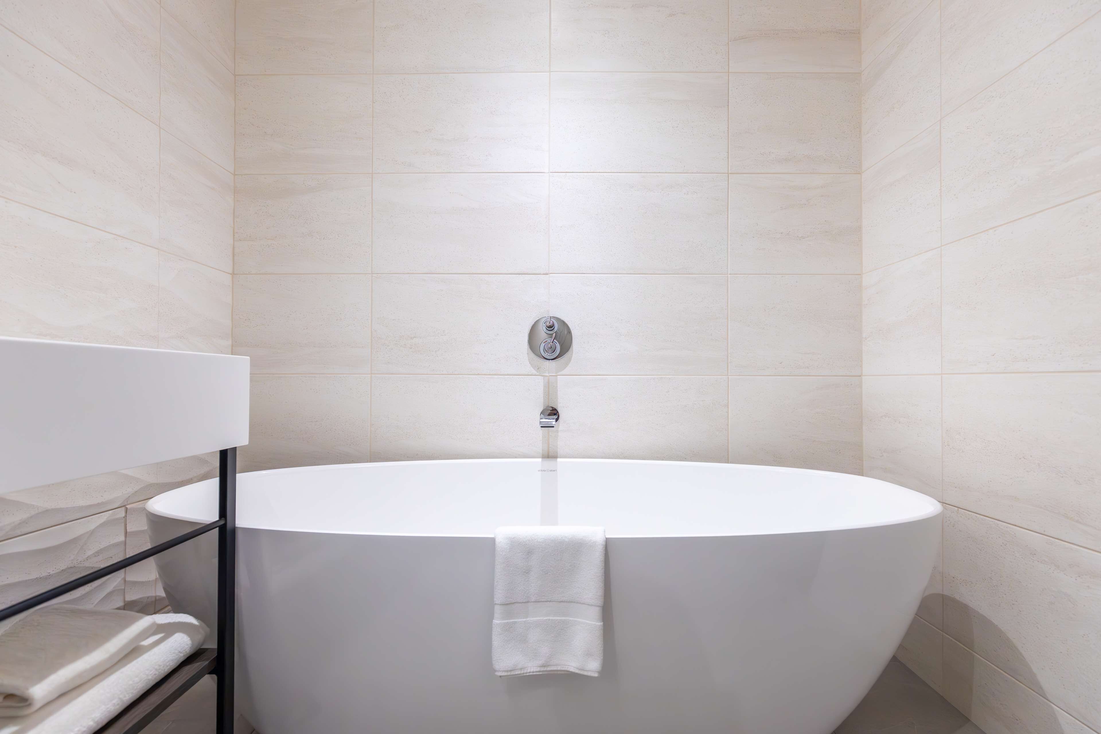 Een moderne badkamer met een vrijstaand bad, een glazen plank met handdoeken en een wandmengkraan.
