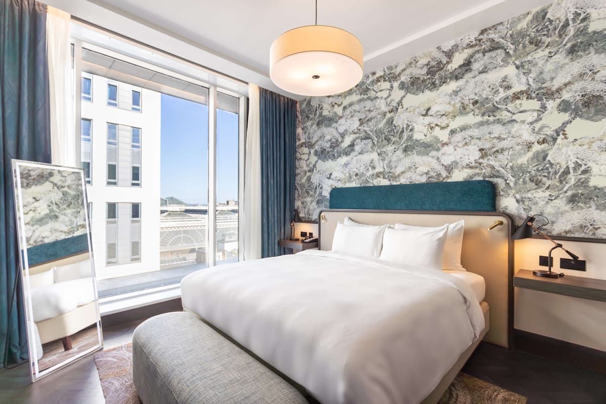 A modern hotel room with a large bed, a view of the city, and a marble-patterned wall.