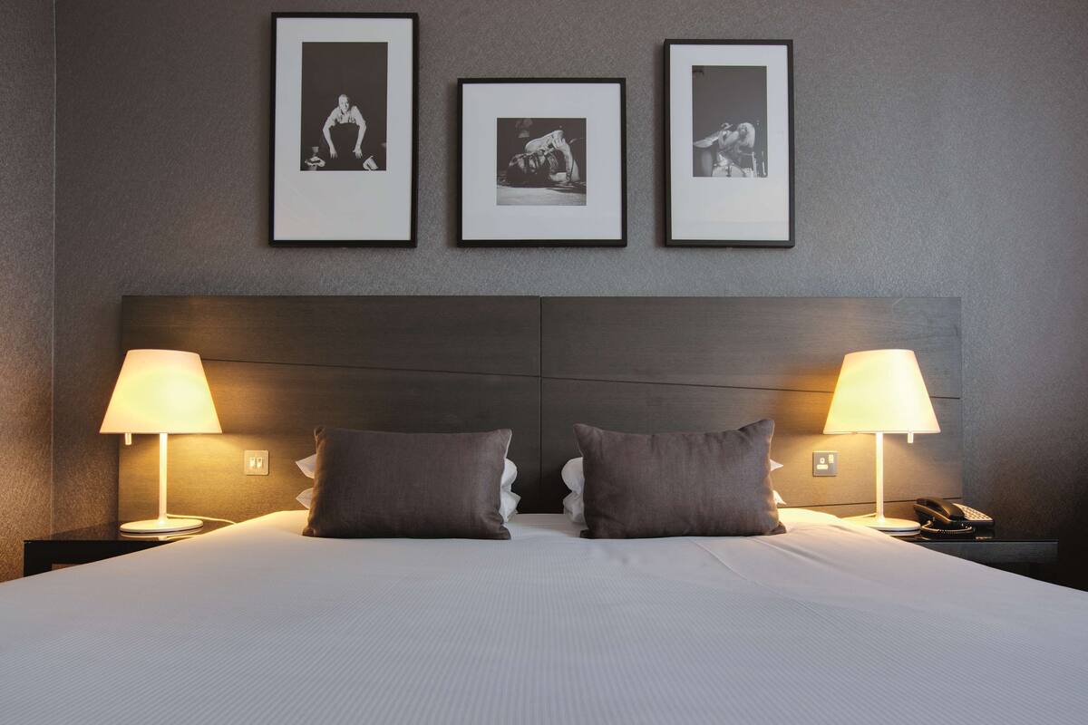 A hotel room with a large bed, two bedside lamps, a telephone, and three framed pictures on the wall.