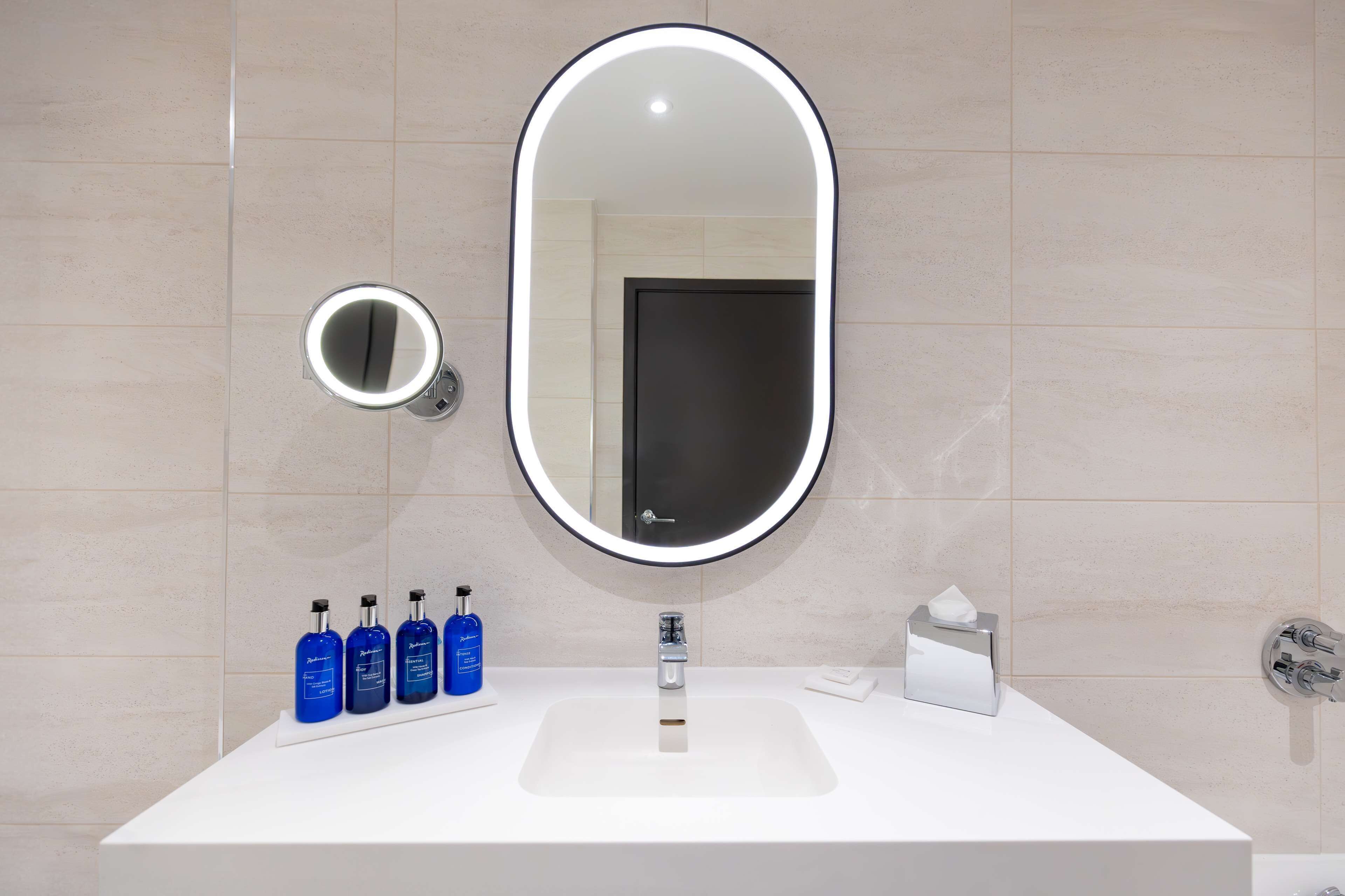 Un baño con un lavabo blanco, un espejo ovalado grande con luz y cuatro botellas azules sobre la encimera.