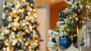 Un sapin de Noël décoré de guirlandes lumineuses et d’ornements, un escalier orné d’une guirlande, et un arrière-plan flou avec un mur et une porte.