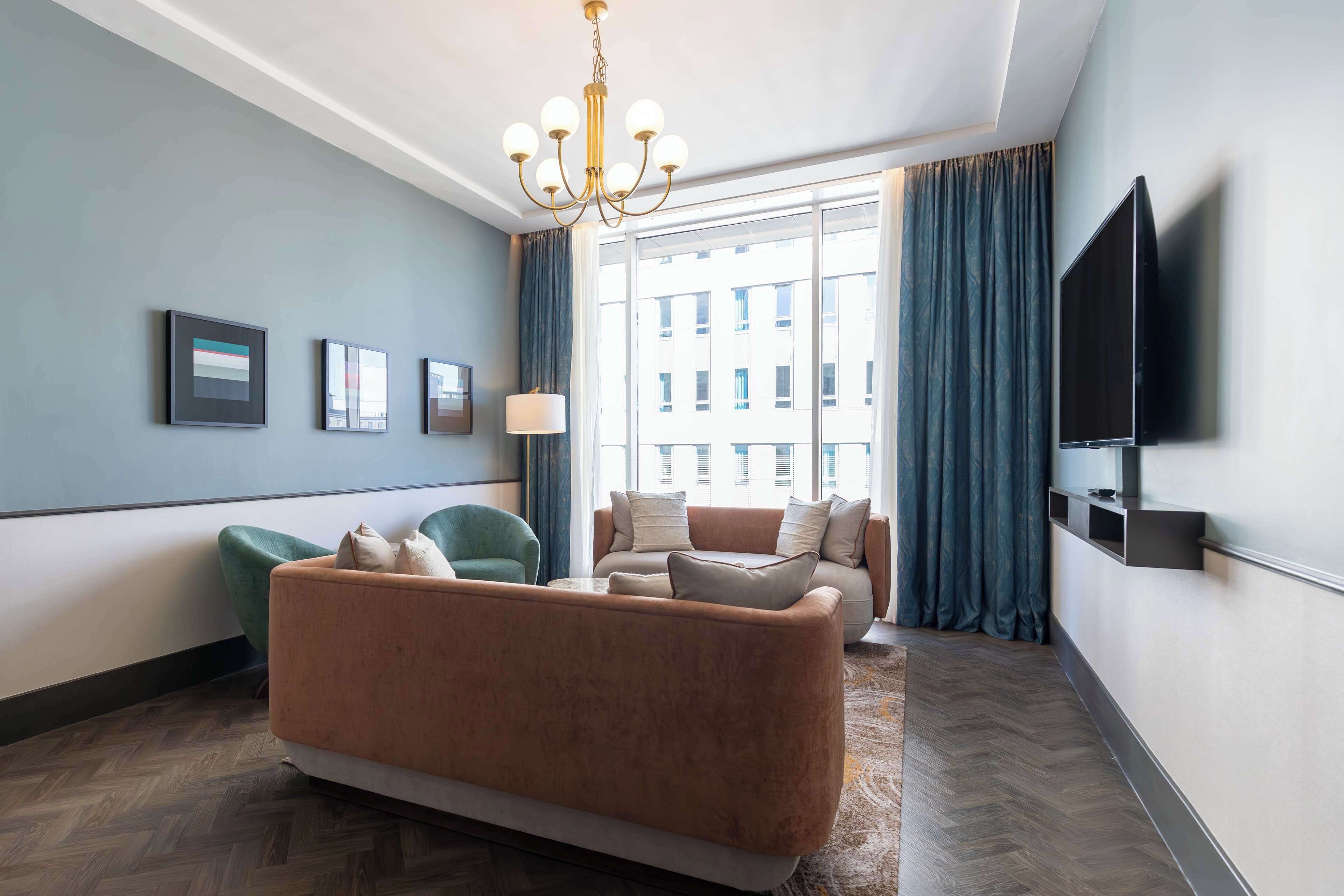 A modern hotel room with a large sofa, a smaller armchair, a flat-screen TV, and a large window with curtains.