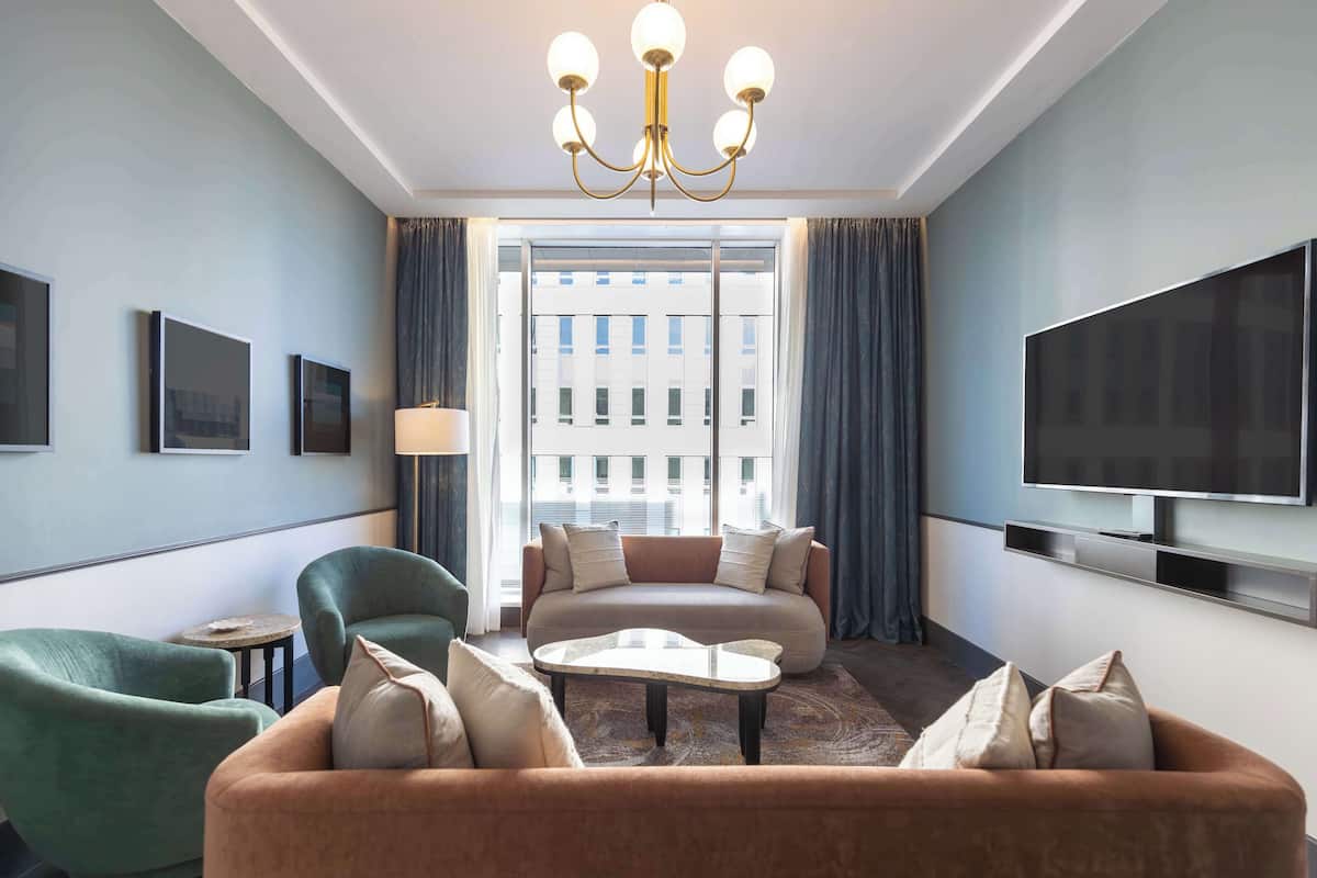 A modern hotel room with a sofa, armchairs, a coffee table, a TV, and a large window with curtains.
