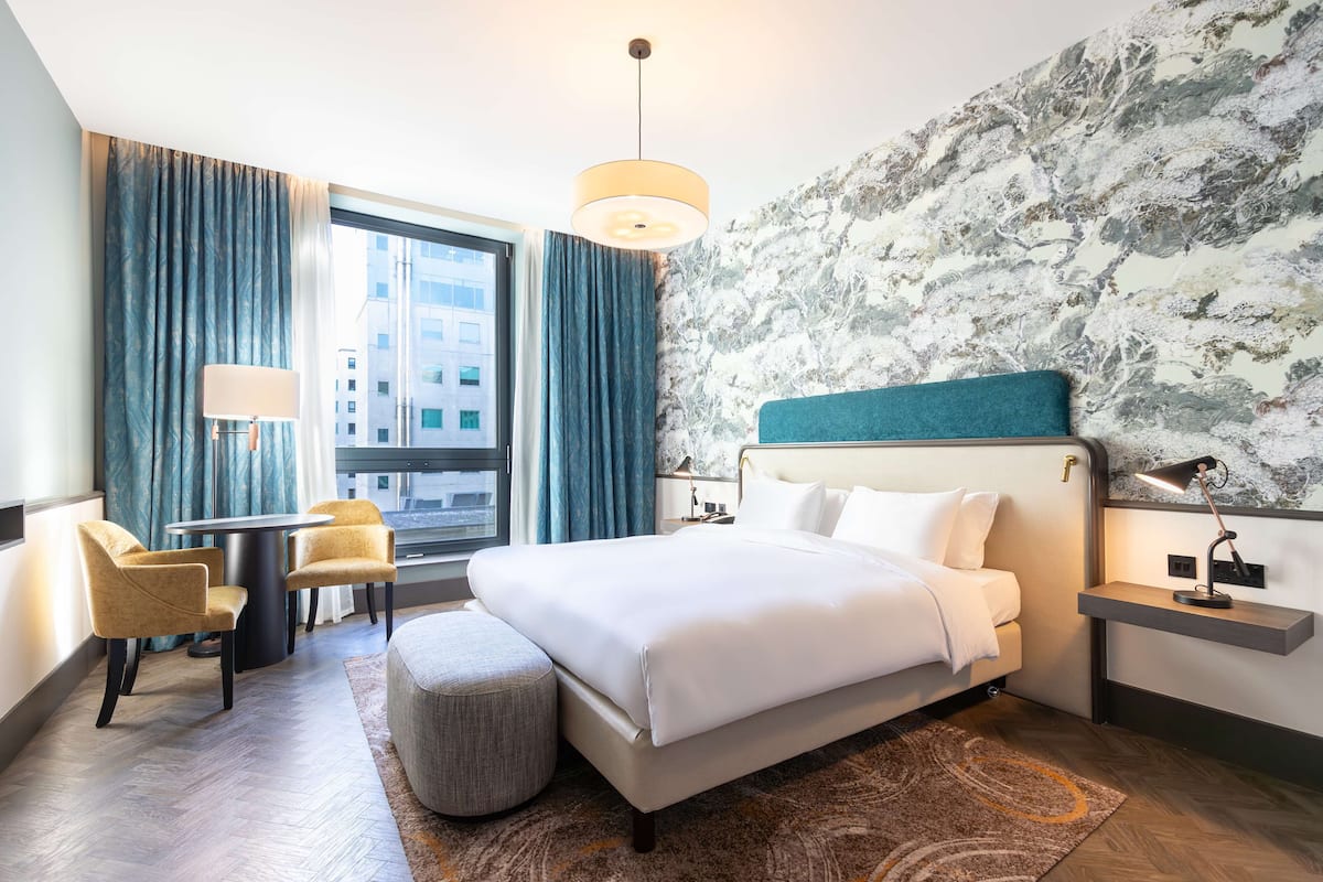 A modern hotel room with a large bed, a desk, two chairs, a round ottoman, a nightstand with a lamp, and a view of the city through large windows.