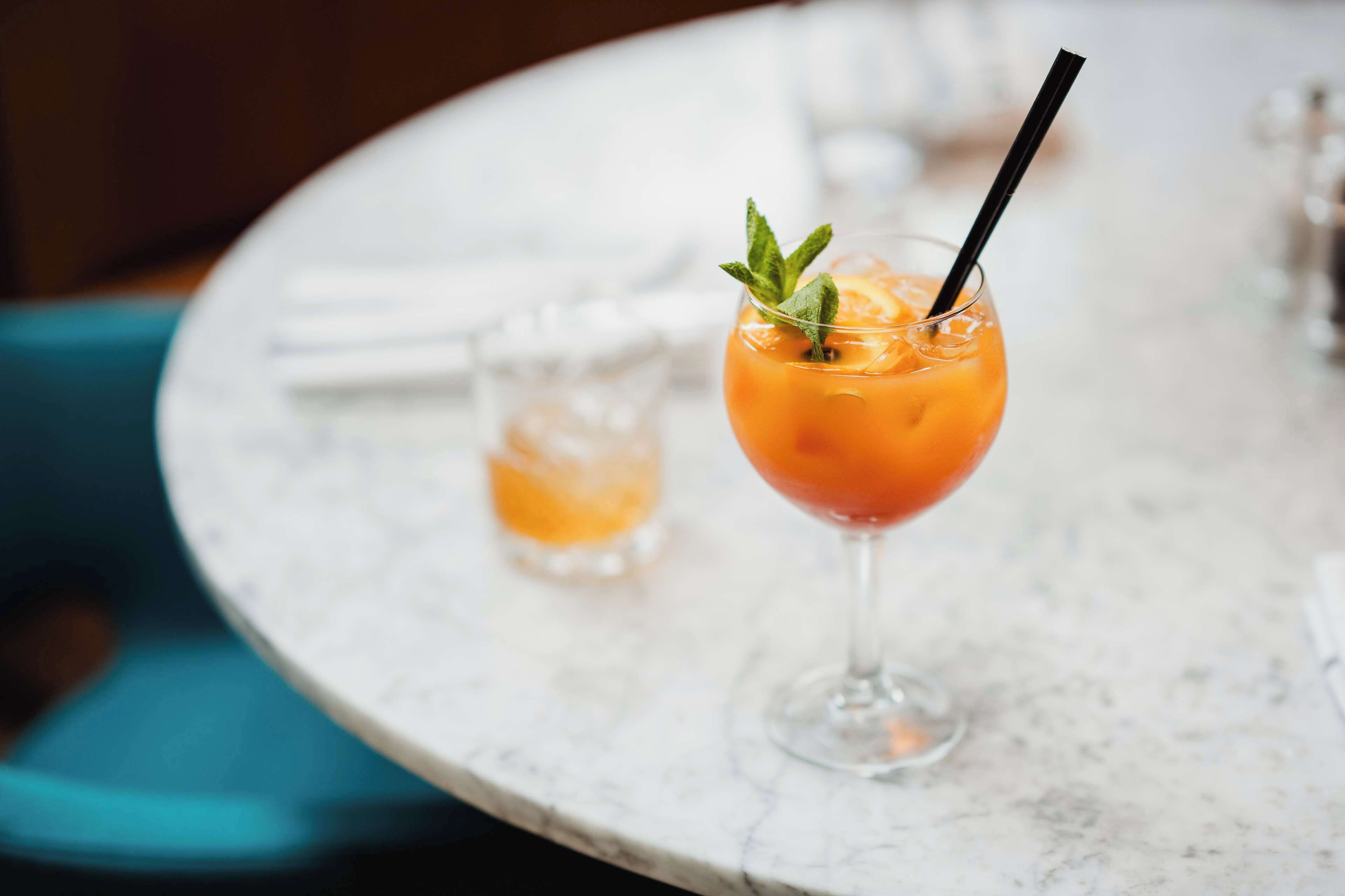 A round table with two glasses of orange-colored drinks, one with a mint garnish and a black straw.