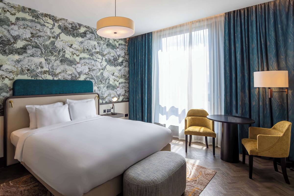 A hotel room with a large bed, a desk, two chairs, a lamp, and a view of the outdoors through sheer curtains.