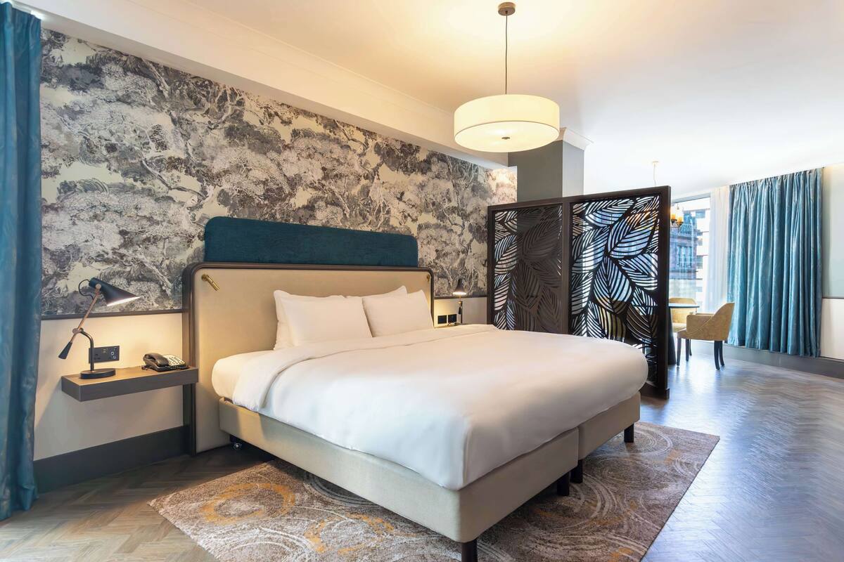 A modern hotel room with a large bed, a nightstand with a lamp, a desk with a chair, and a decorative wall with a leaf pattern.