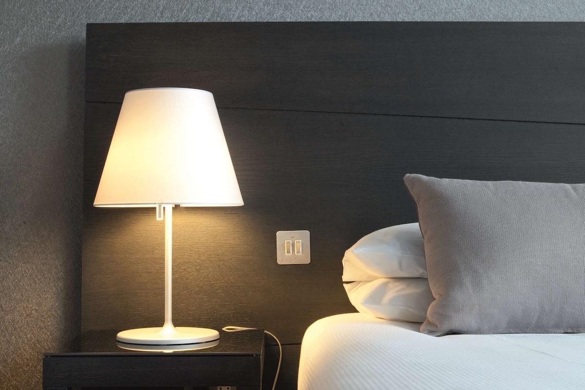 A neatly made bed with a white comforter and pillows, a bedside table with a lamp, and framed pictures on the wall.