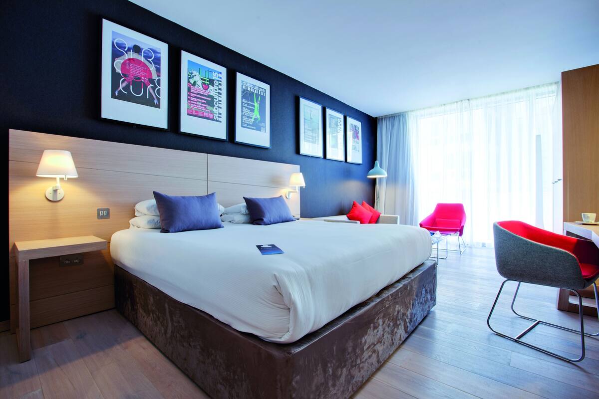 A modern hotel room with a large bed, bedside tables, a desk, a chair, and framed pictures on the wall.