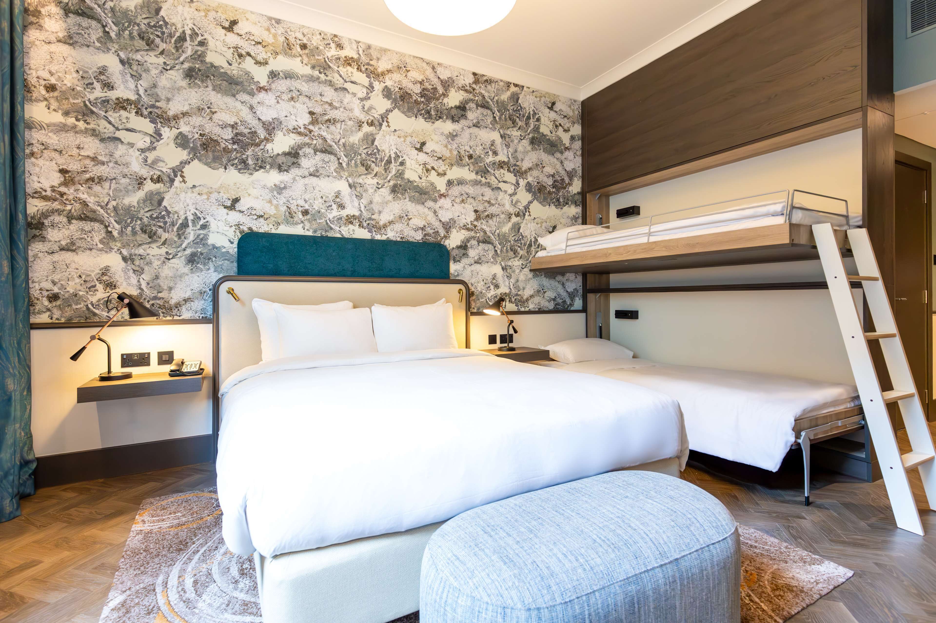 Un dormitorio con una cama litera, una mesita de noche con una lámpara y un otoman redondo.