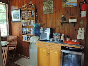 A cozy cabin interior with wooden walls, a coffee machine, a bookshelf filled with books and nautical decorations, and a small dining area. - Shorelands Guest Resort and Cottages (Kennebunk)