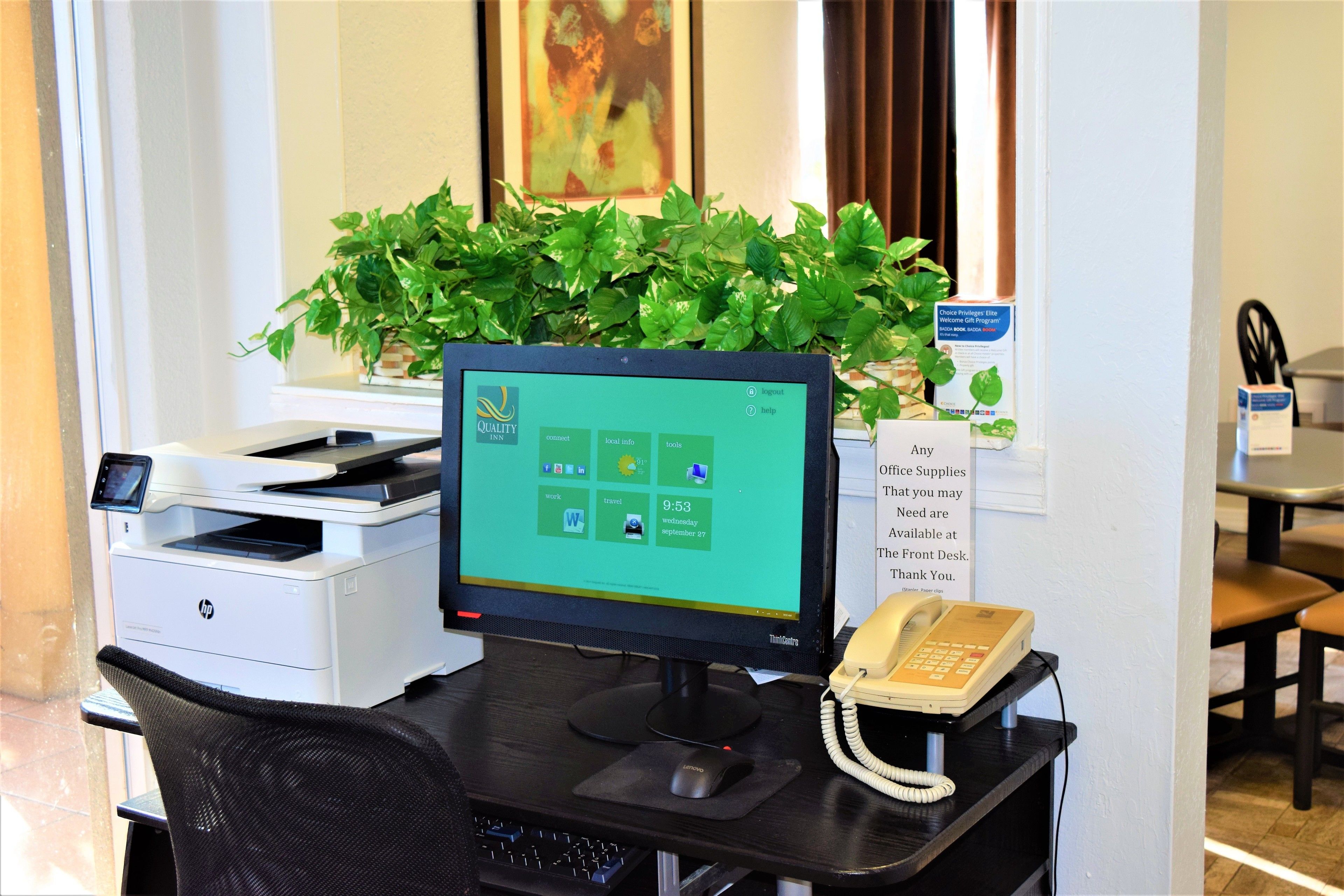 A reception desk with a computer monitor, printer, and telephone. There is a plant on the desk, and a sign about available service supplies.