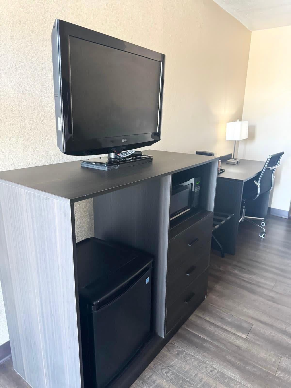 Una habitación de hotel moderna con un televisor de pantalla plana montado en un escritorio de madera oscura, un pequeño mueble de almacenamiento negro, una lámpara de escritorio y una silla de oficina negra.