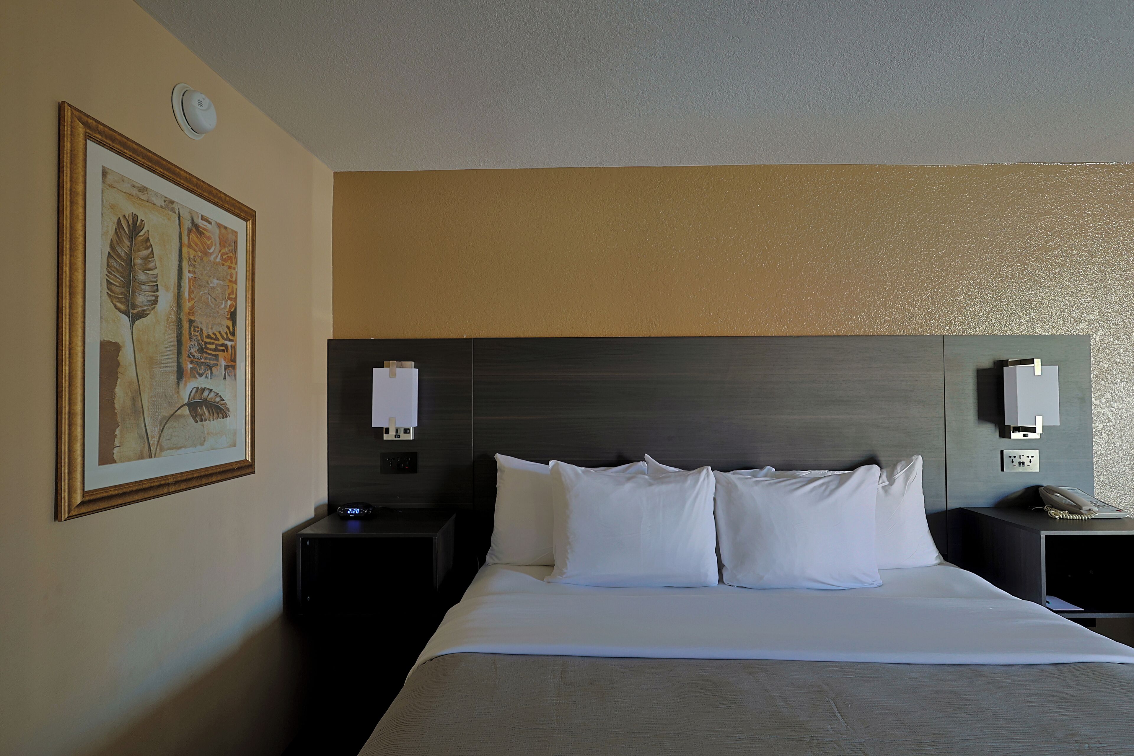 Una habitación de hotel con una cama grande, dos mesitas de noche, un velador, un reloj de pared y una obra de arte enmarcada.