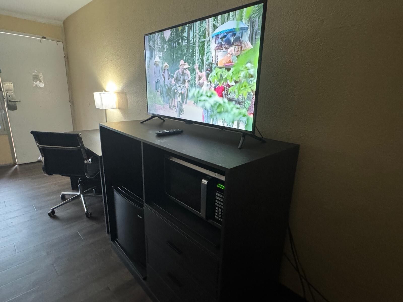 A television on a black stand in a room with a wooden floor, a chair, and a door.