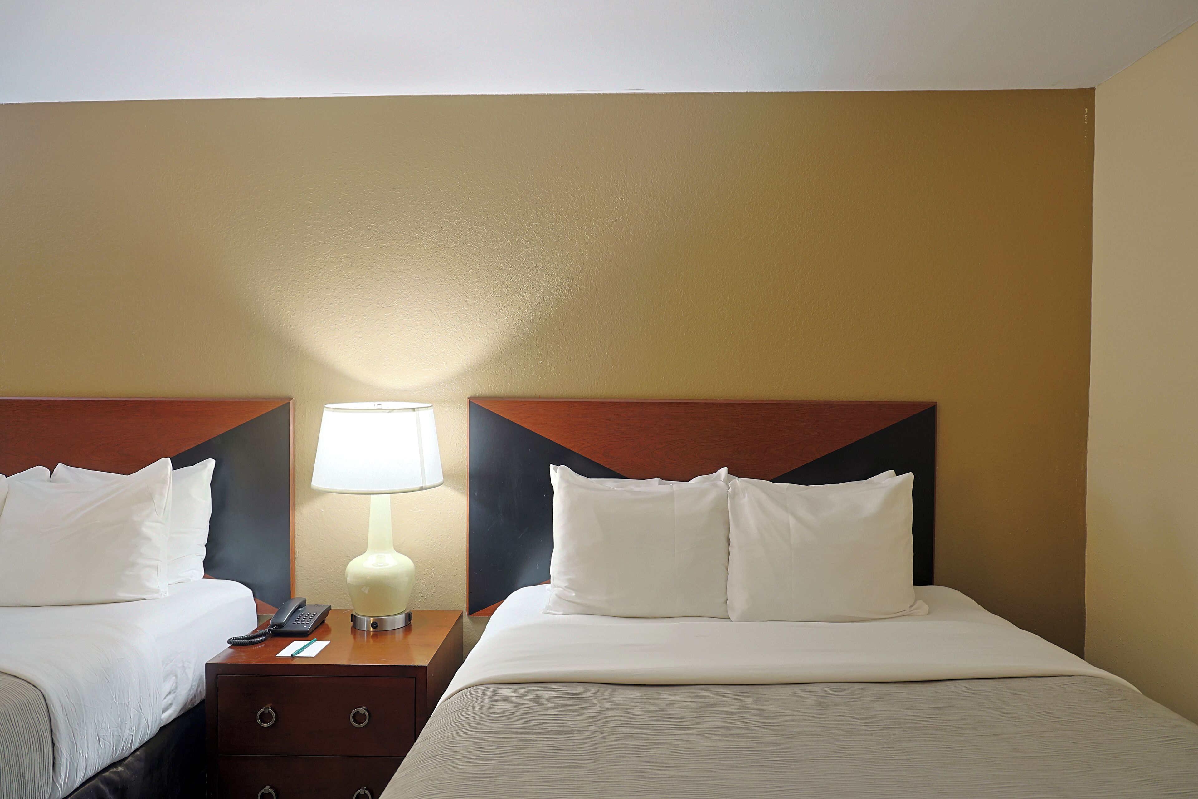 A hotel room with two beds, each with white bedding and pillows, a nightstand with a lamp, and a telephone.