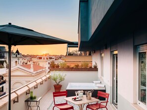 A balcony with a table, chairs, a potted plant, and a view of the cityscape at sunset.