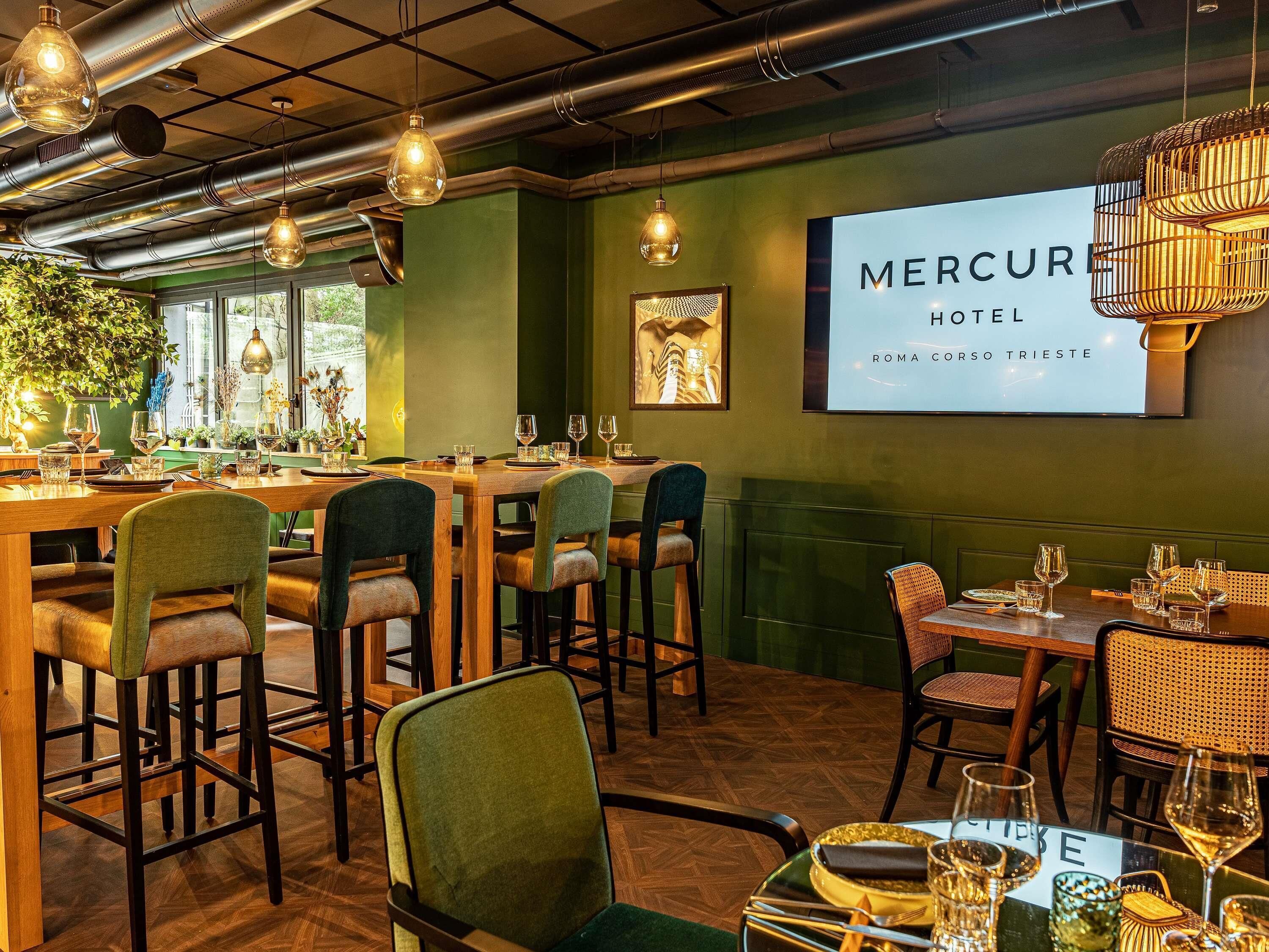 A restaurant with tables set for dining, a projection screen displaying 'MERCURE HOTEL ROMA CORSO TRIESTE', and hanging industrial-style lights.