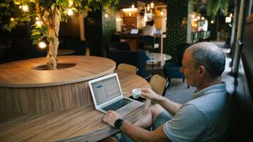 A man sitting at a round wooden table with a laptop and a cup, in a cafe with a tree and hanging lights in the background.