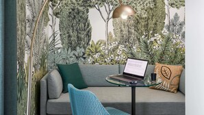 A room with a sofa, a glass table with a laptop, a lamp, and a wall with a nature-themed mural.