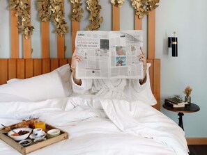 A person reading a newspaper on a bed with white linens, a tray of breakfast items, and a wooden headboard with decorative elements.