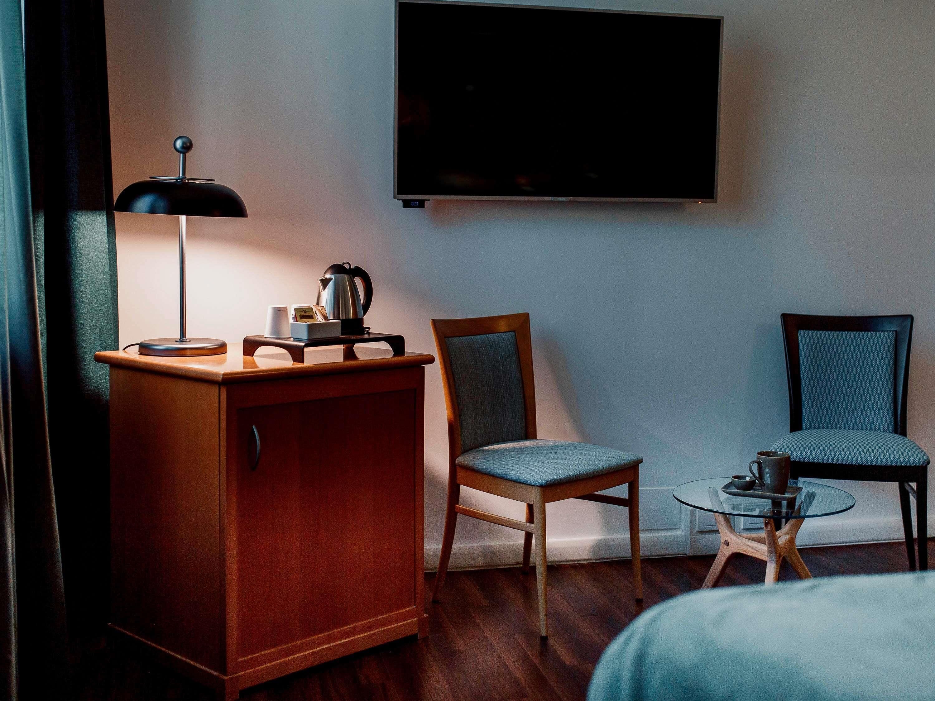Ett hotellrum med ett träskåp, en stol, ett litet bord med en vattenkokare, en tv som är monterad på väggen och en säng som syns delvis.
