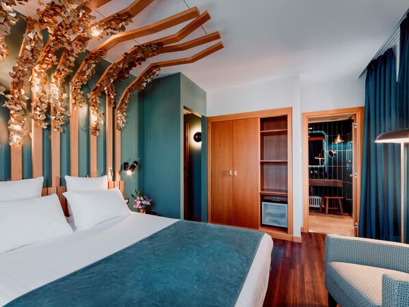 A modern hotel room with a bed, a wooden wardrobe, a seating area, and a bathroom visible through an open door.