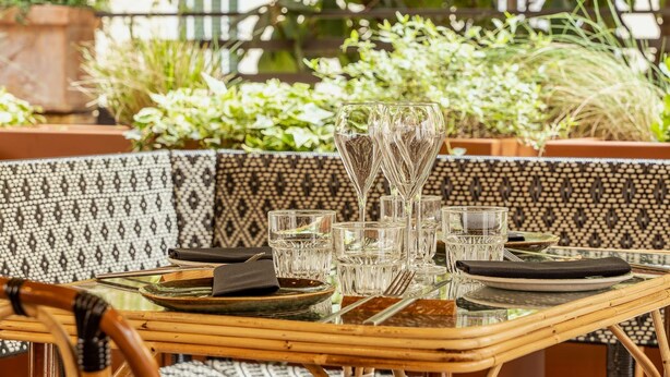 A table set with clear glasses, plates, and cutlery, surrounded by wicker chairs and a patterned tablecloth.