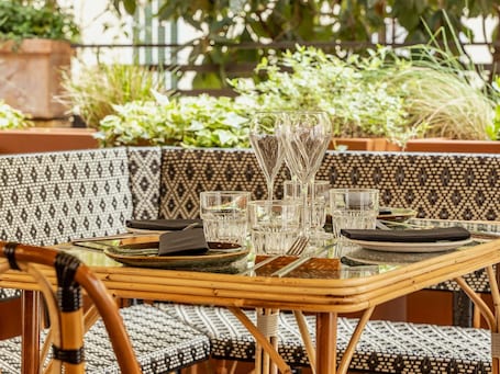 A table set with clear glasses, plates, and cutlery, surrounded by wicker furniture and potted plants.. Mercure Roma Corso Trieste