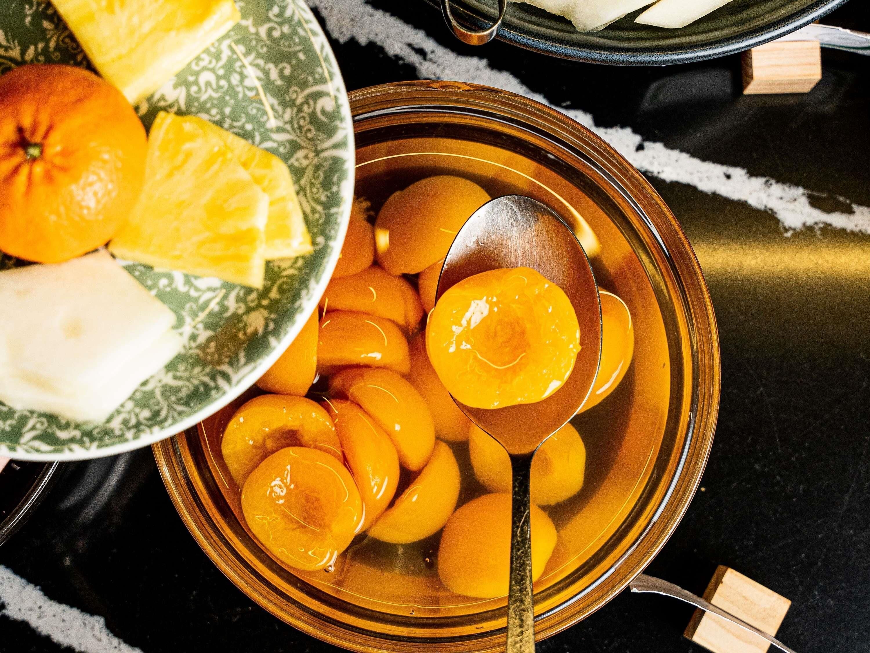 Un bol rempli de tranches de pêches, avec une cuillère à côté, une assiette avec une orange et un objet blanc, et un bol à motifs en arrière-plan.