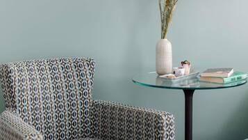 A patterned armchair, a glass table with a vase and books, and a plain wall.