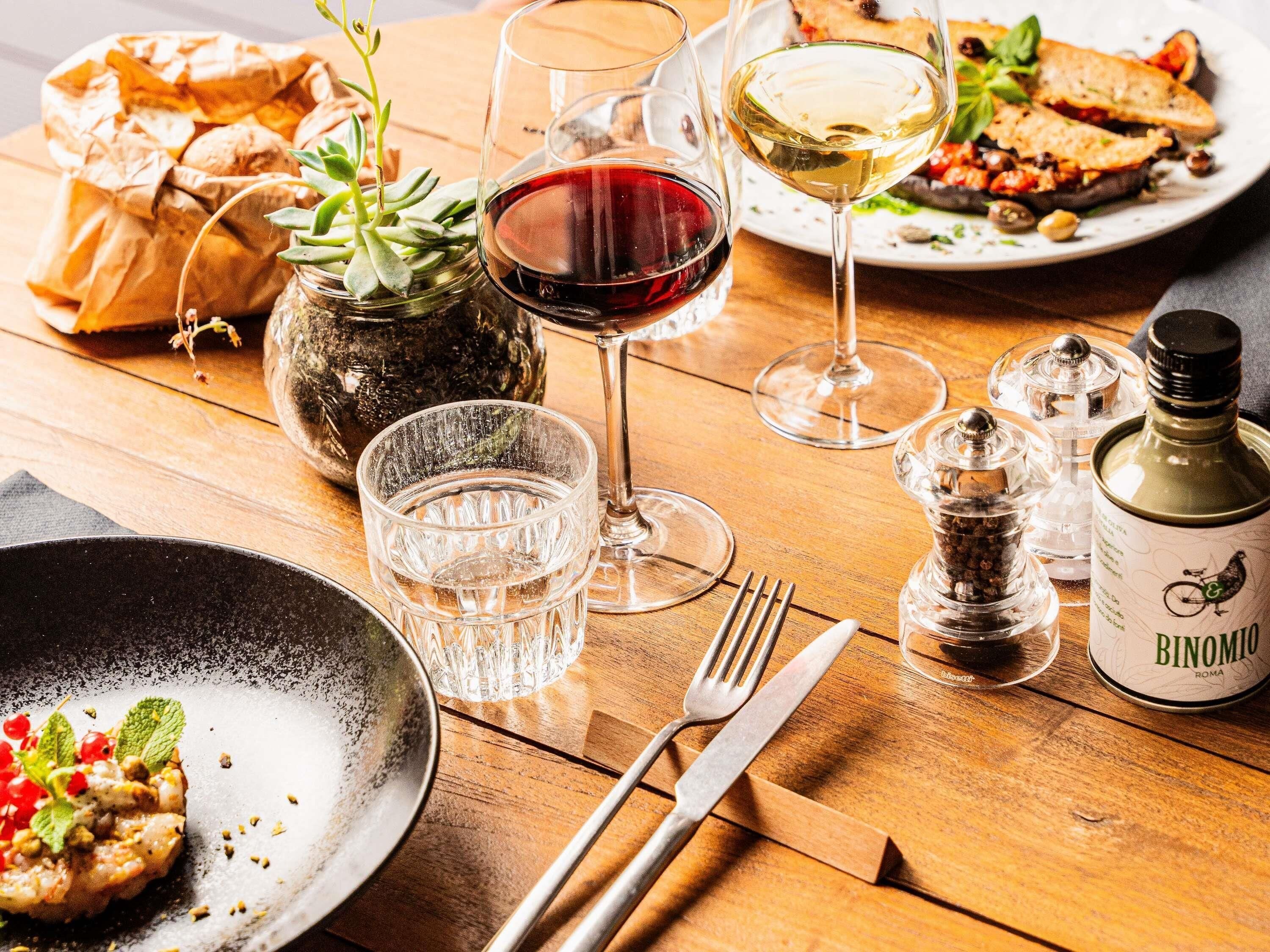 A dining table with plates of food, glasses of wine, cutlery, and a bottle of Binomio wine.