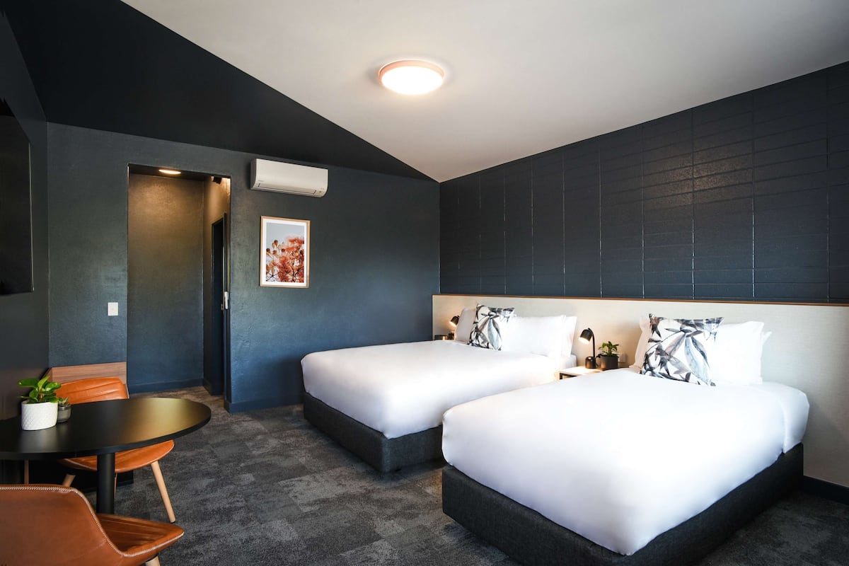 Une chambre d’hôtel avec deux lits, une petite table et une chaise. Il y a un luminaire au plafond et un appareil de climatisation.