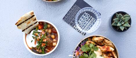 A variety of dishes including a bowl of salad, a bowl of soup, a plate of chicken, and a box of fries. There are also two small potted plants and a coaster with text.